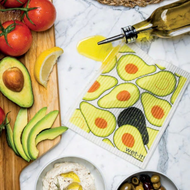 Avocado patterned Wet-it towel laying on a counter soaking up spilled olive oil next to an olive oil bottle and freshly sliced fruits and veggies