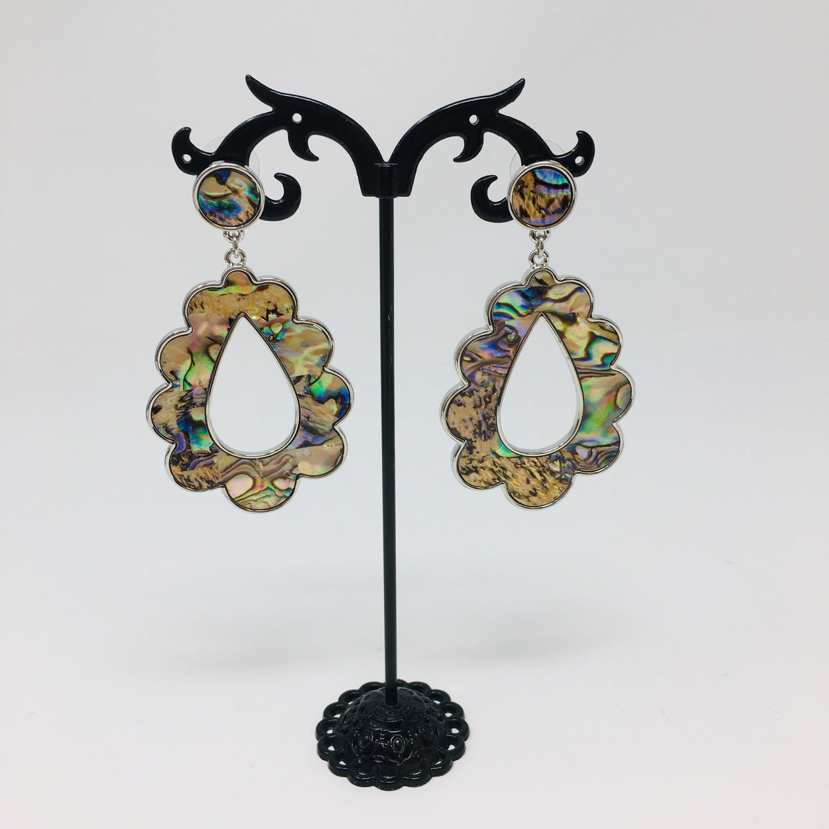 Scalloped teardrop shaped silver tone earrings with abalone shell inserts shown hanging from a black earring stand on a white background