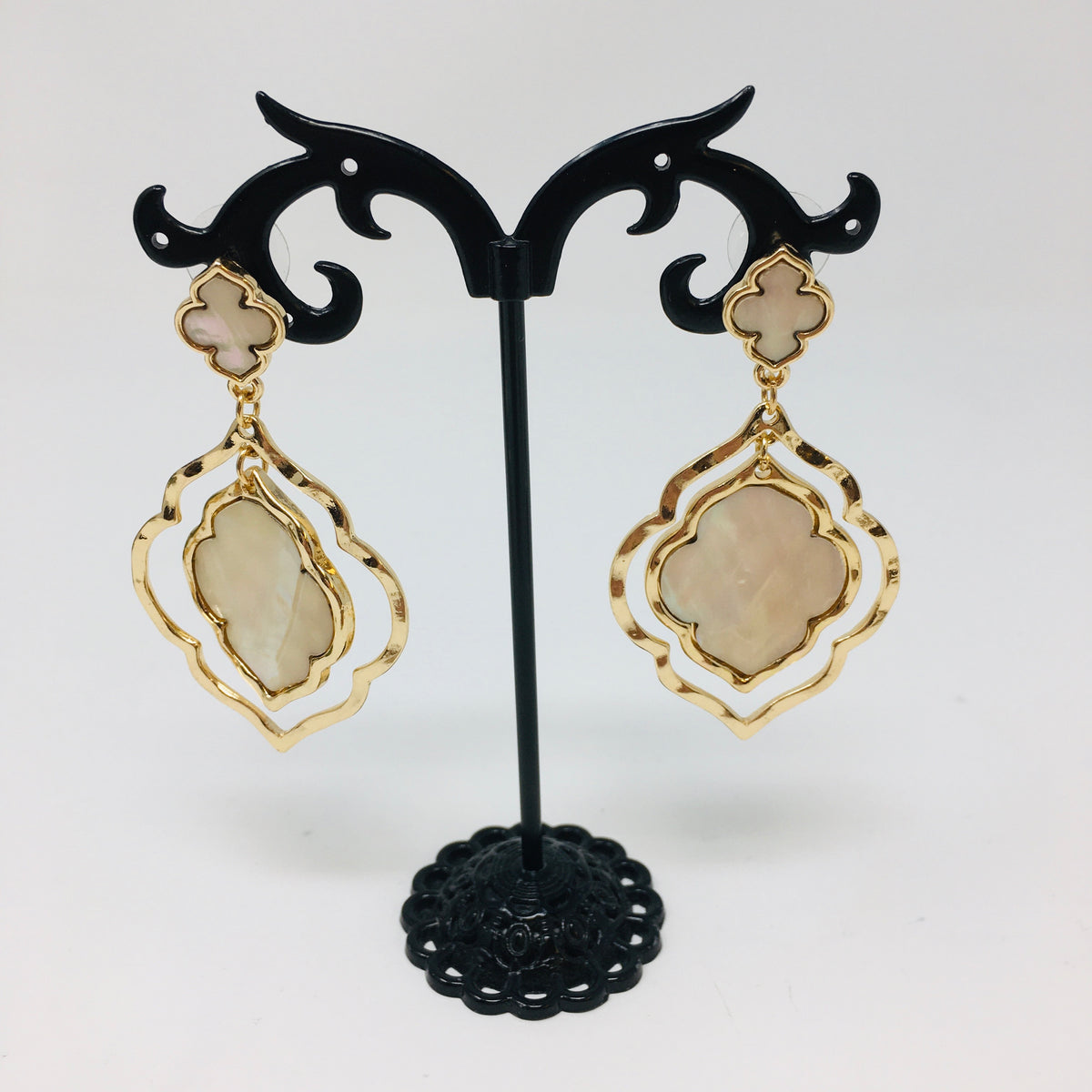 double layer arabesque shaped gold tone earrings with mother of pearl inserts shown hanging from a black earring stand on a white background